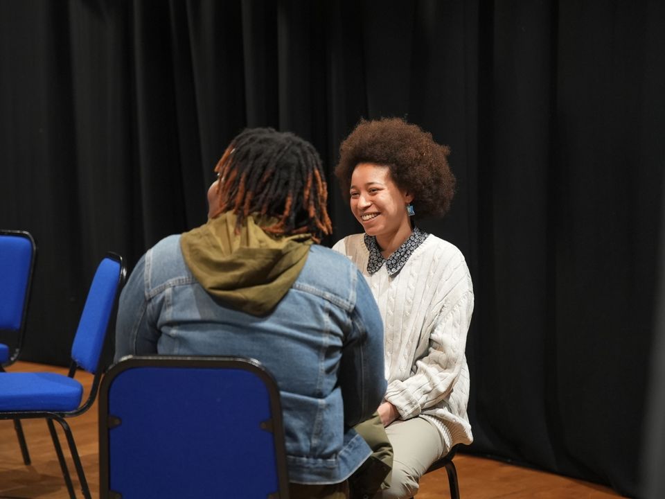 Photo of two people sat on blue chairs in front of a black curtain, with one person looking at and smiling at the other who has their back turned to the camera and is looking to the side