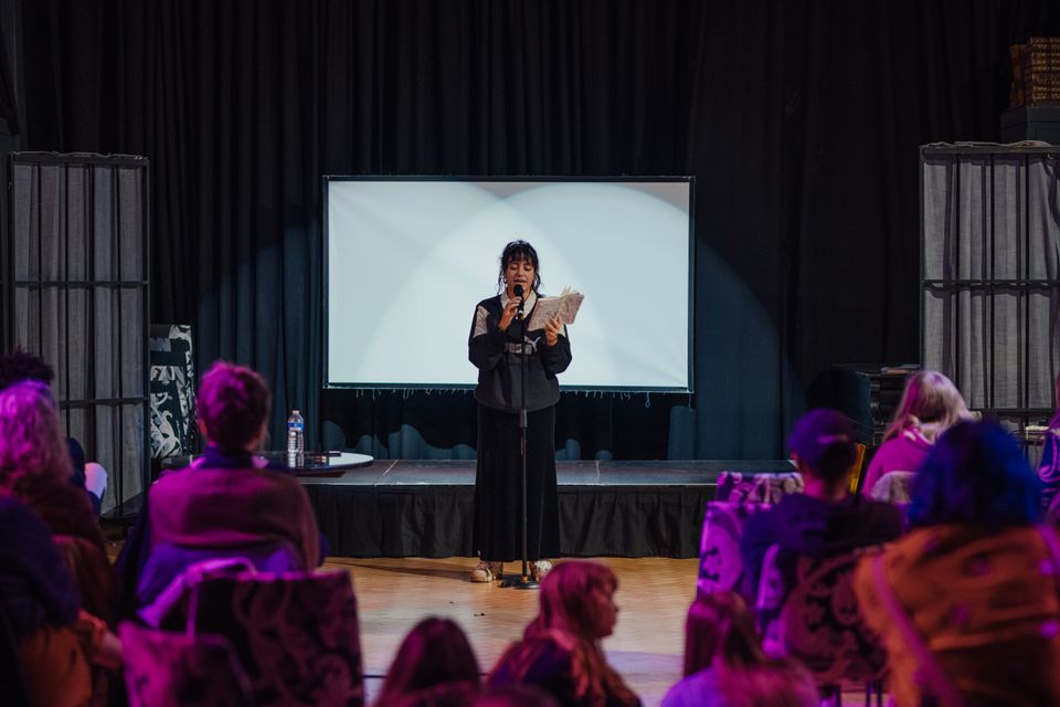 Photo of a person stood in front of a dimly lit seated audience reading from a book and speaking into a microphone. There is a stage and a white screen behind the person speaking, and they are lit up by a spot light.