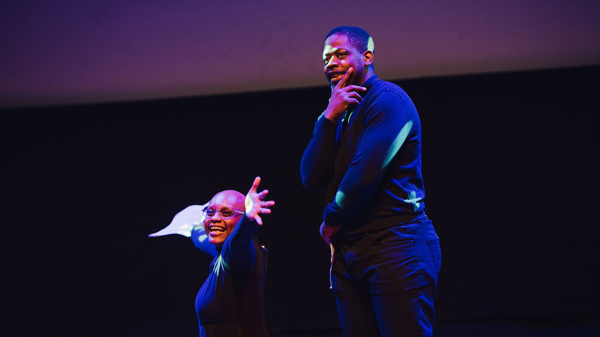 Photo of two people smiling and performing in front of a black and purple background, with one kneeling with their arms outstretched and the other standing with one of their hands on their chin